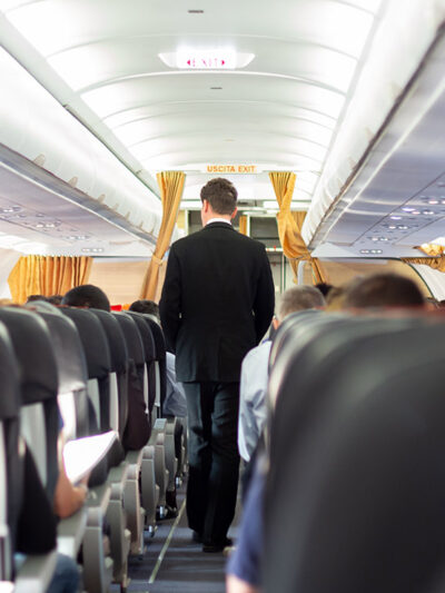 A flight attendant wearing a suit walking through the aisle of a plane.