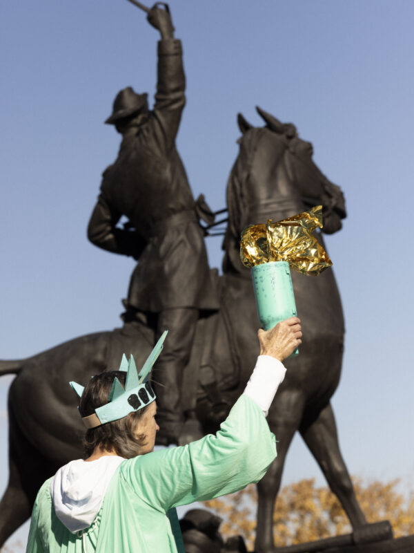 Someone dressed as the Statue of Liberty standing in front of a statue of a man on a horse