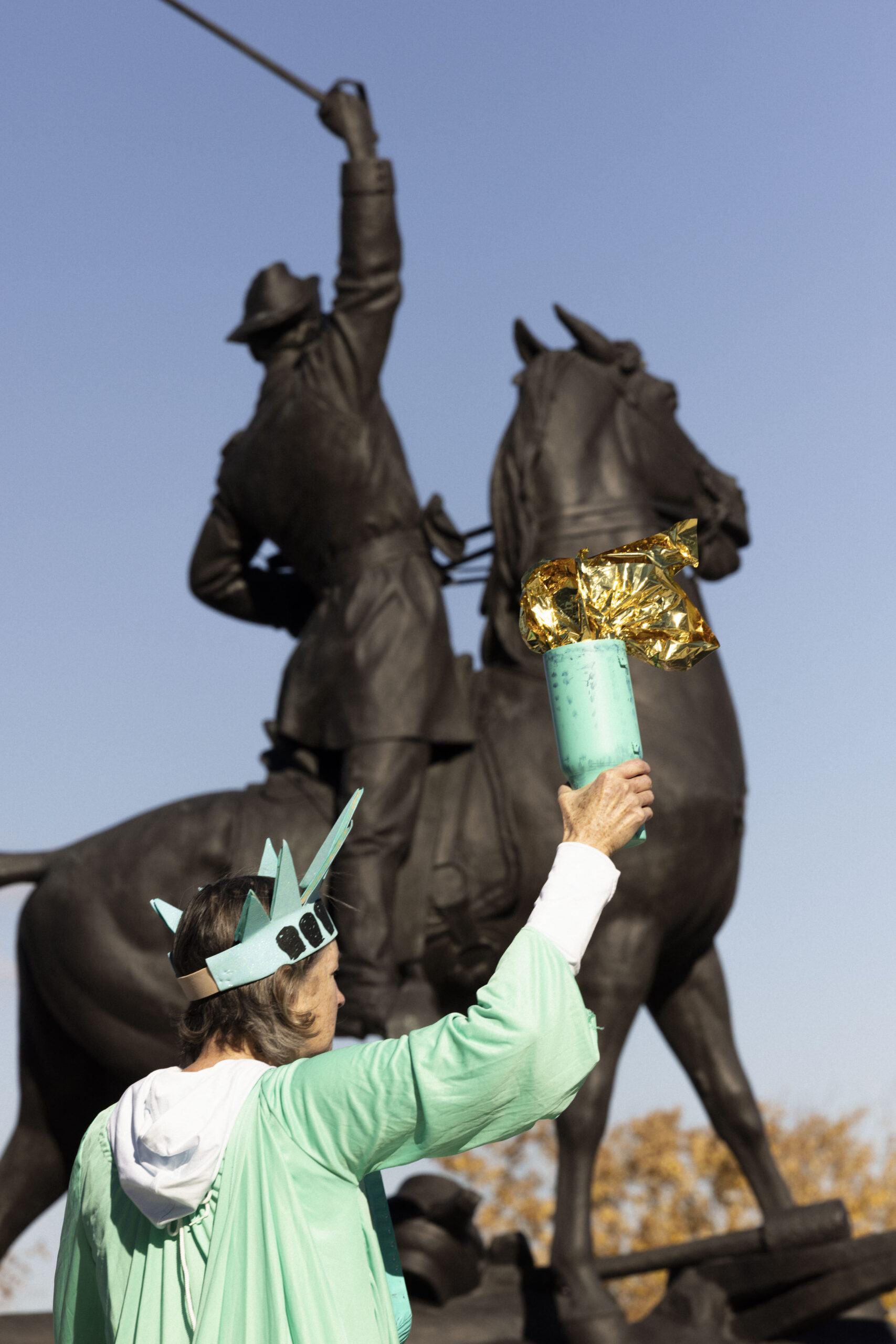 Someone dressed as the Statue of Liberty standing in front of a statue of a man on a horse