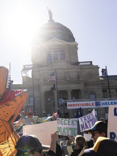 A photo of people rallying in front of the Montana State Capitol, including someone dressed in an inflatable dinosaur costume