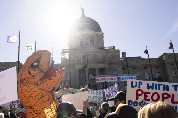 A photo of people rallying in front of the Montana State Capitol, including someone dressed in an inflatable dinosaur costume