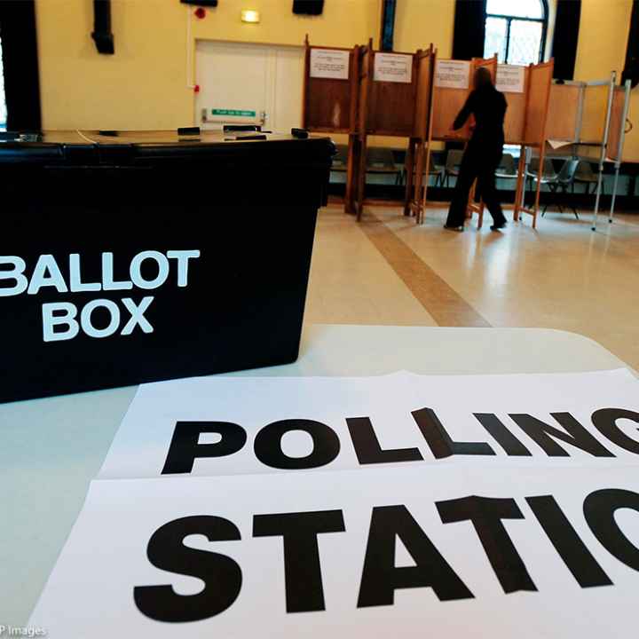 In the foreground, a black box with the words "BALLOT BOX" rests on a sign reading "POLLING STATION" at a polling station, while in the background a person in silhouette is at a voting booth.